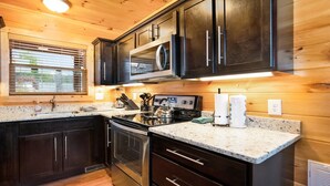 A modern kitchen with dark cabinets, granite countertops, stainless steel appliances, and a window with blinds. - Cozy 2bedroom cabin near Island / Dollywood at Pigeon Forge  with Panormic Views (Sevierville)