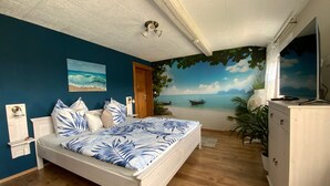 Ein Schlafzimmer mit einem Bett, einer Kommode, einem Deckenventilator, einem Strandbild und einem Wandgemälde mit einer Strandansicht.