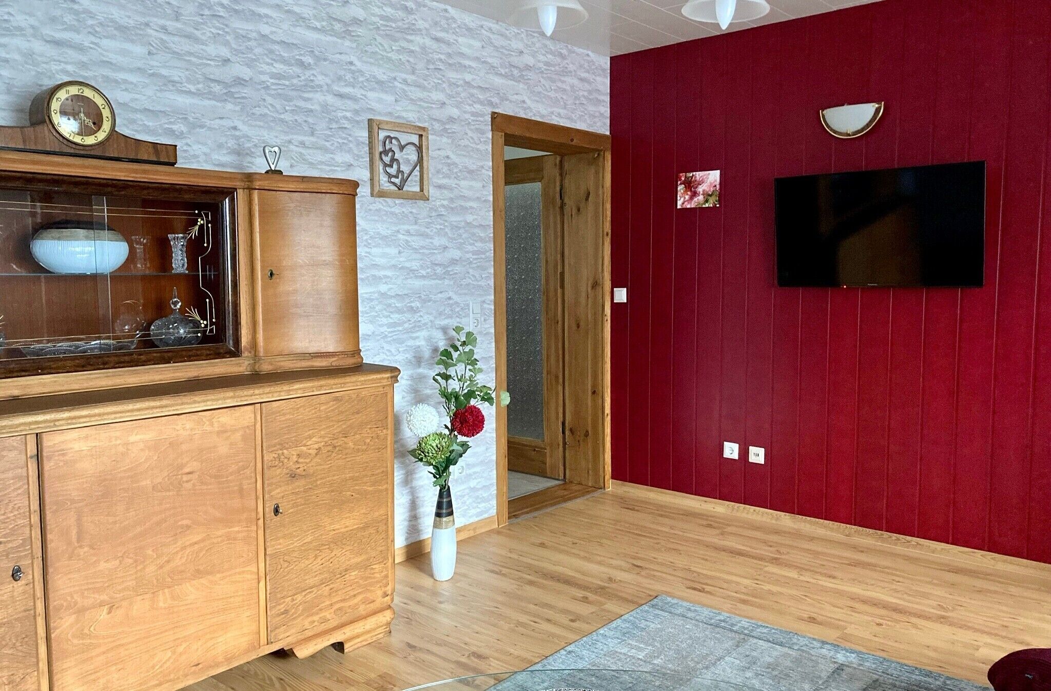 A room with a wooden cabinet, a vase with flowers, a red wall, a television, and a clock.