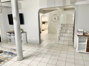 Um interior amplo com piso de azulejos, uma escadaria e uma área de cozinha visível através de um arco.