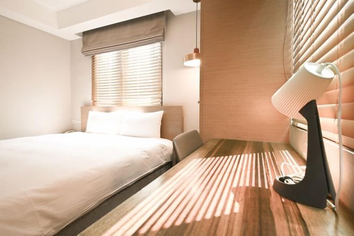 Una camera d'albergo con un letto, un comodino, una lampada e una finestra con persiane.