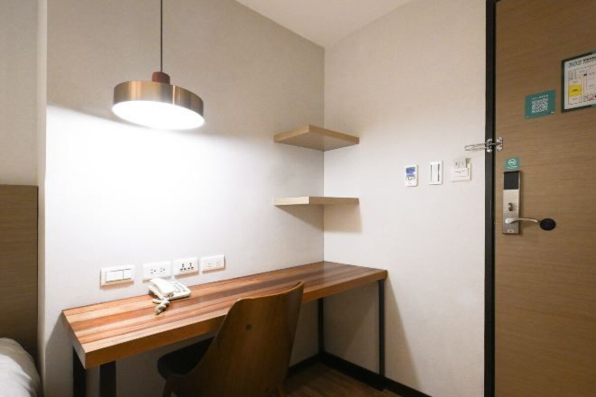 A compact room with a wooden desk, a chair, a hanging light fixture, a door with a lock, and a wall-mounted electrical outlet.