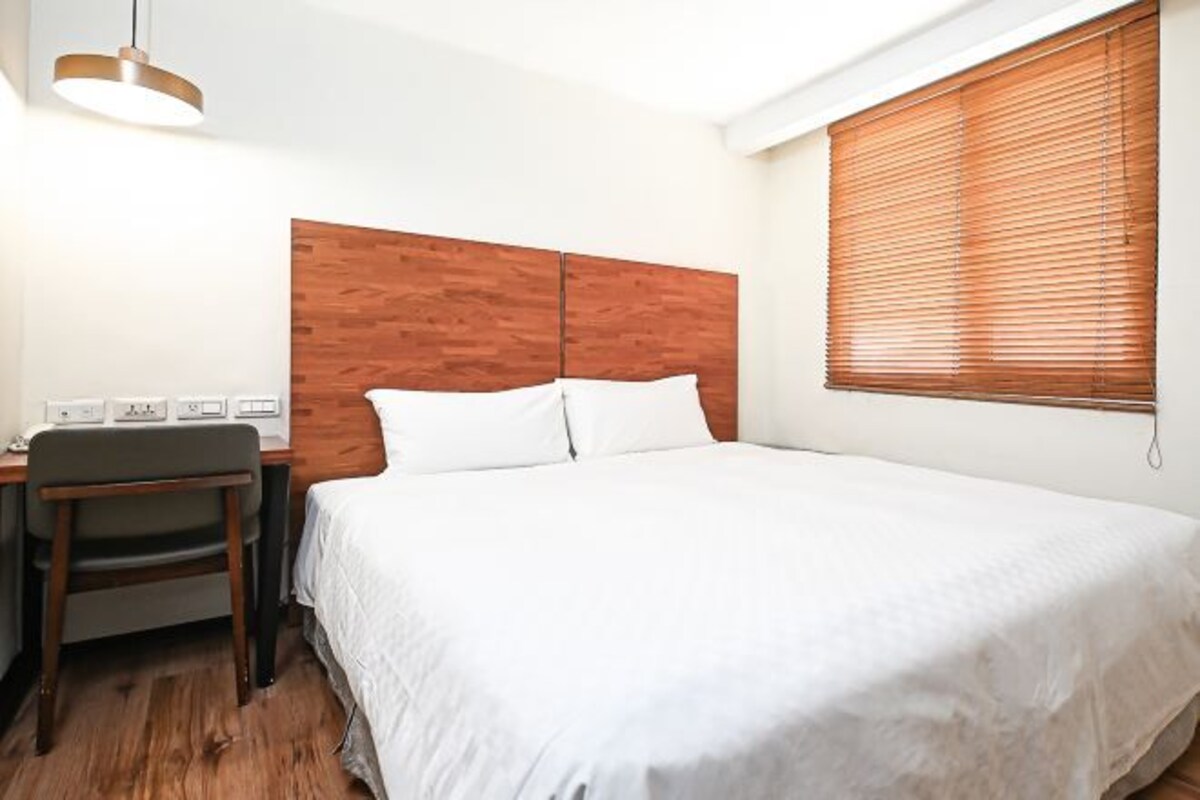 A hotel room with a wooden headboard, a bed with white linens, a desk with a chair, and a window with blinds.