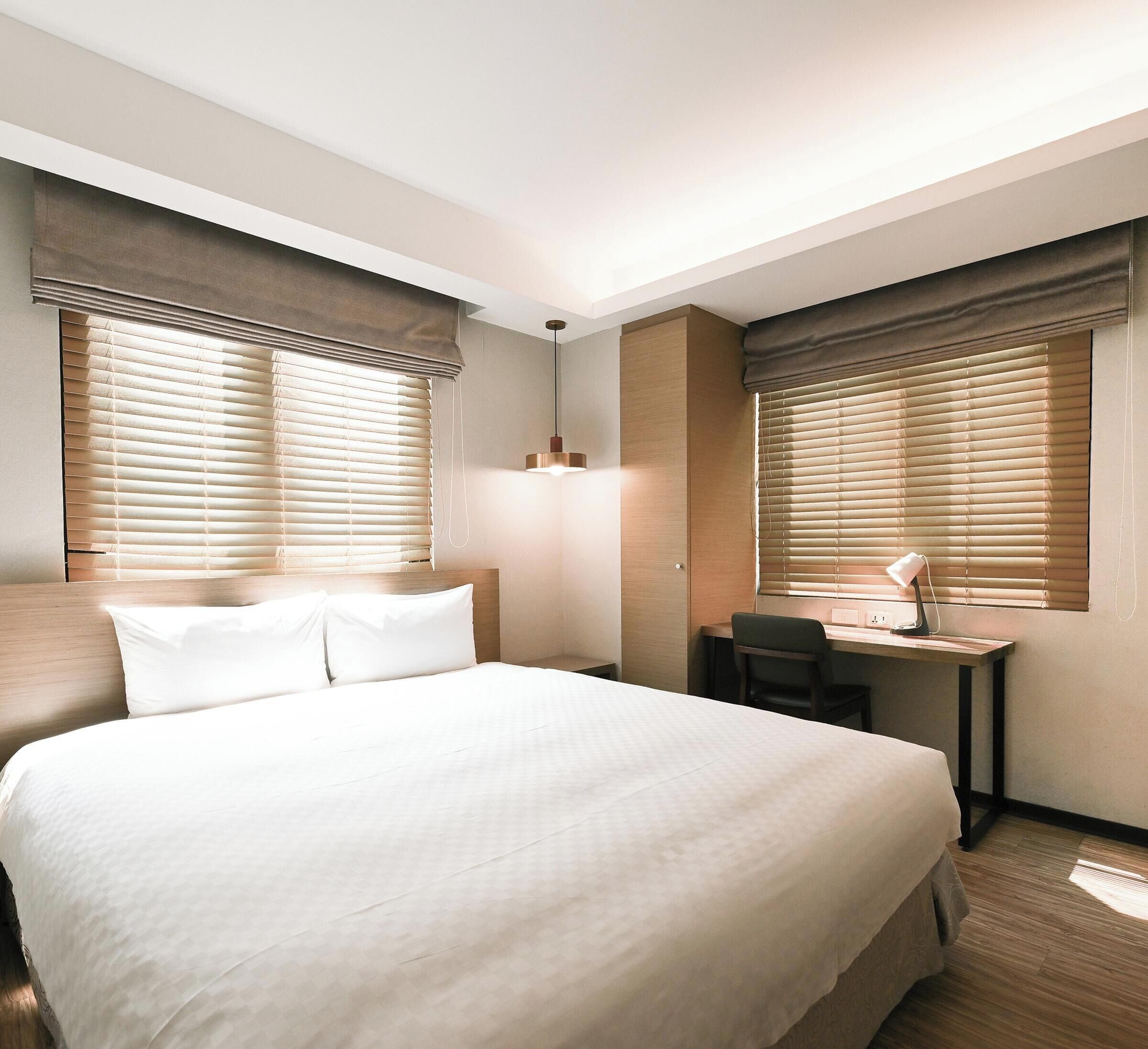 Habitación de hotel con una cama grande, un escritorio con silla y dos ventanas con persianas.