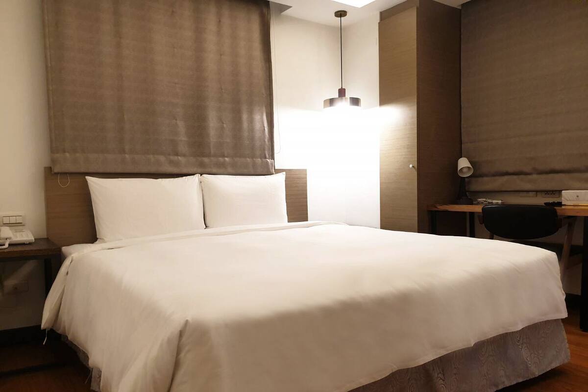 Une chambre d’hôtel avec un grand lit, un bureau avec une chaise, une lampe et une fenêtre avec des rideaux.