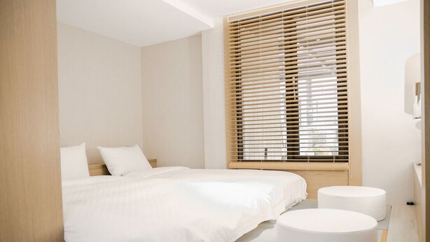 A modern bedroom with a large bed, two white round ottomans, and a window with horizontal blinds. - CHECK inn Express Taipei Station (Taipei)