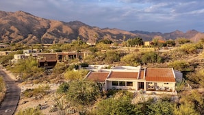 A residential area with houses surrounded by mountains, featuring a paved road and sparse vegetation.