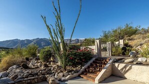 A garden with a stone pathway leading to a metal gate, surrounded by desert vegetation and mountains in the background.