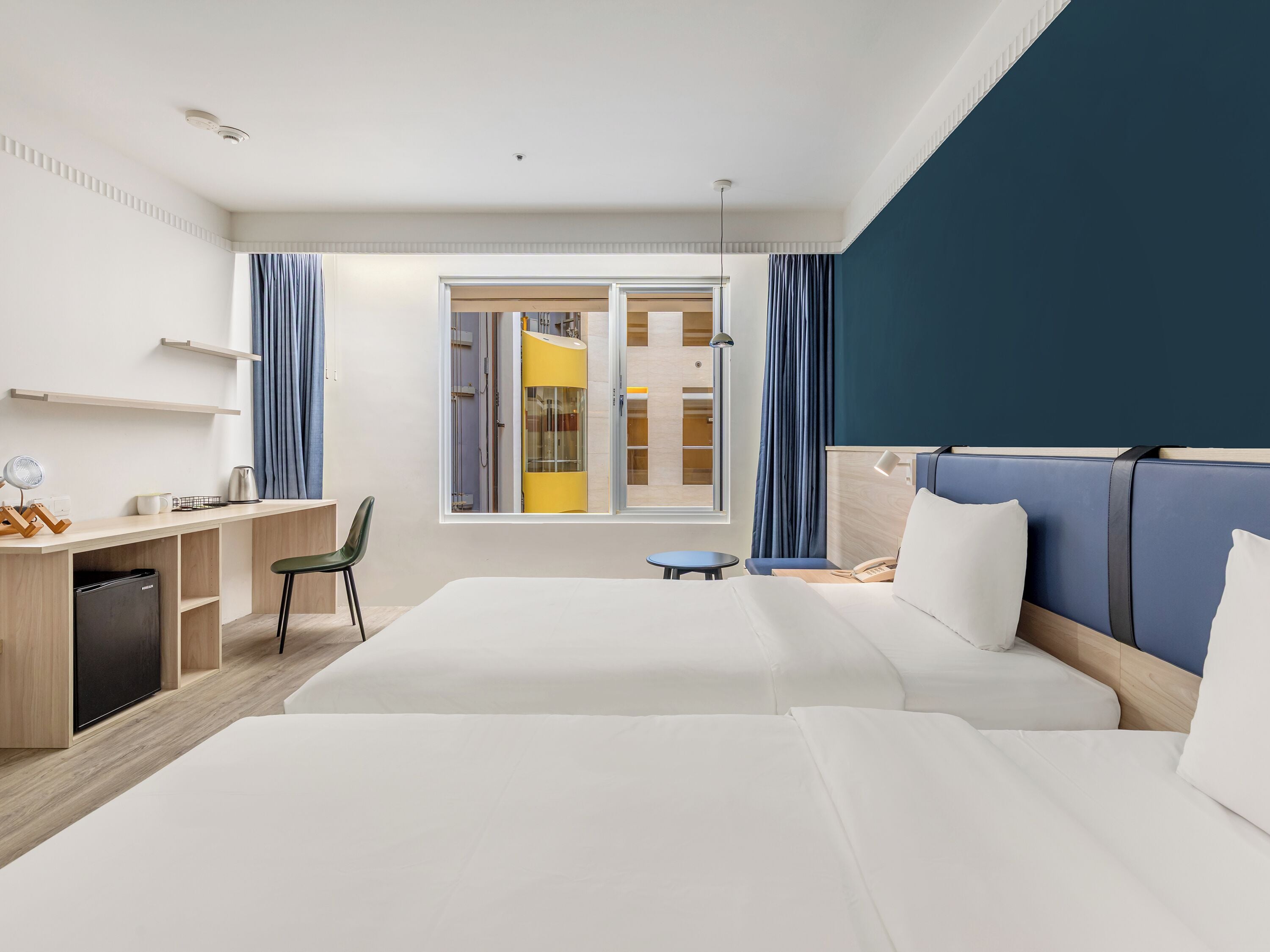 A hotel room with a bed, desk, chair, and a view of a yellow elevator.