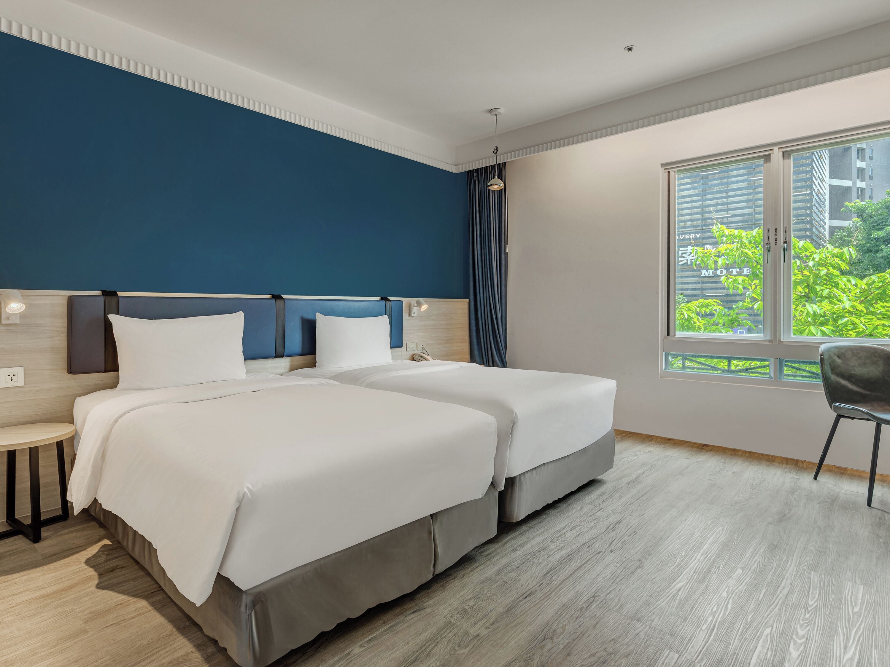 Habitación de hotel con dos camas, una mesita de noche, una silla, ventana con vistas a zonas verdes y una lámpara en el techo.