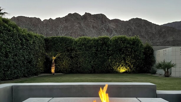 Una estructura de piedra rectangular con fuego, rodeada por un seto cuidado y con montañas al fondo.
