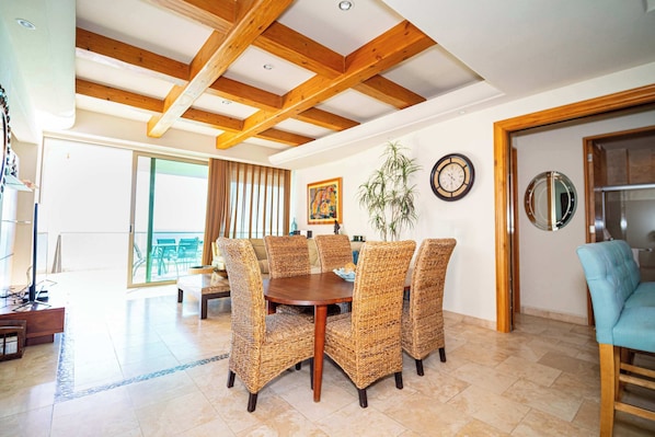 A dining area with a round table and wicker chairs, wooden beams on the ceiling, and a view to an outdoor patio.