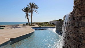 A pool with a waterfall feature, palm trees, and a view of the ocean. - Beachfront Luxury at Tessoro | 3BR Sleeps 8 | Pool (Puerto Peñasco)