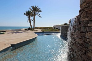 A pool with a waterfall feature, palm trees, and a view of the ocean. - Beachfront Luxury at Tessoro | 3BR Sleeps 8 | Pool (Puerto Peñasco)