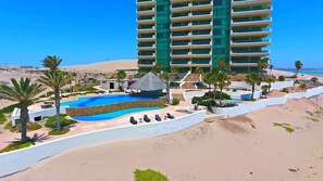 A resort with a swimming pool, palm trees, a beach, and a high-rise building. - Tessoro #1002 at Las Conchas – Elevated Luxury on the Sea of Cortez (Puerto Peñasco)