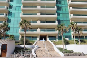 A multi-story hotel with a modern design, featuring a central staircase leading to the entrance, surrounded by palm trees and a landscaped area.