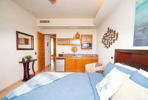 A bedroom with a bed, a bedside table, a nightstand, a chair, a desk, a kitchen area with a microwave, a sink, and a cabinet.