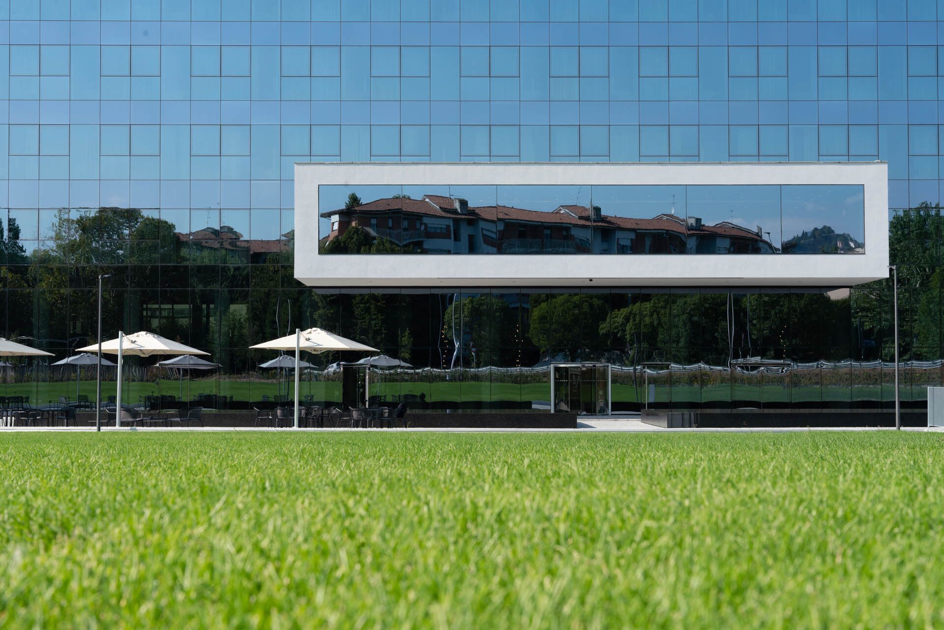 Área externa moderna com mesas e cadeiras sob guarda-sóis, em frente a um edifício com fachada de vidro que reflete as casas próximas.