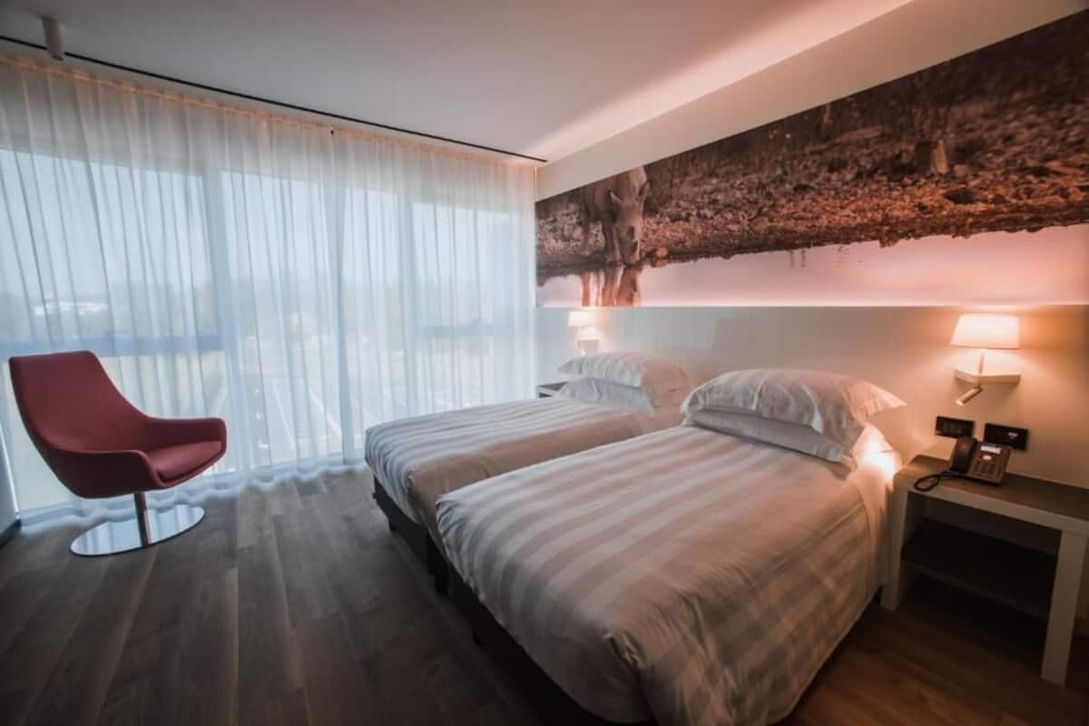 A hotel room with two beds, a red chair, a wall-mounted phone, and a large window with sheer curtains.