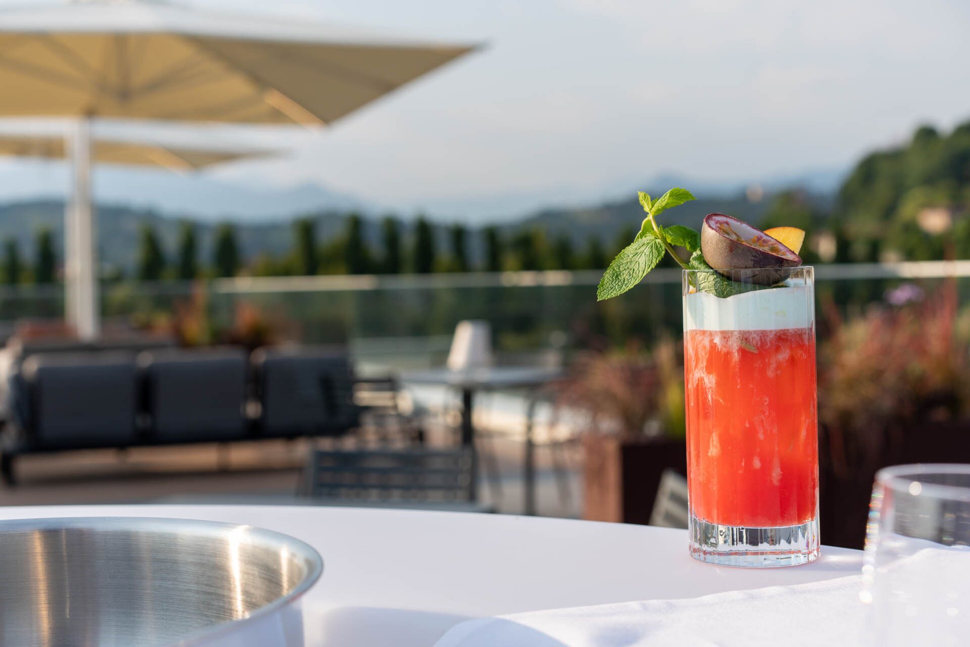 Un grand verre contenant une boisson rouge, agrémenté d’une tranche de fruit et de feuilles de menthe, posé sur une table blanche avec un bol en métal.