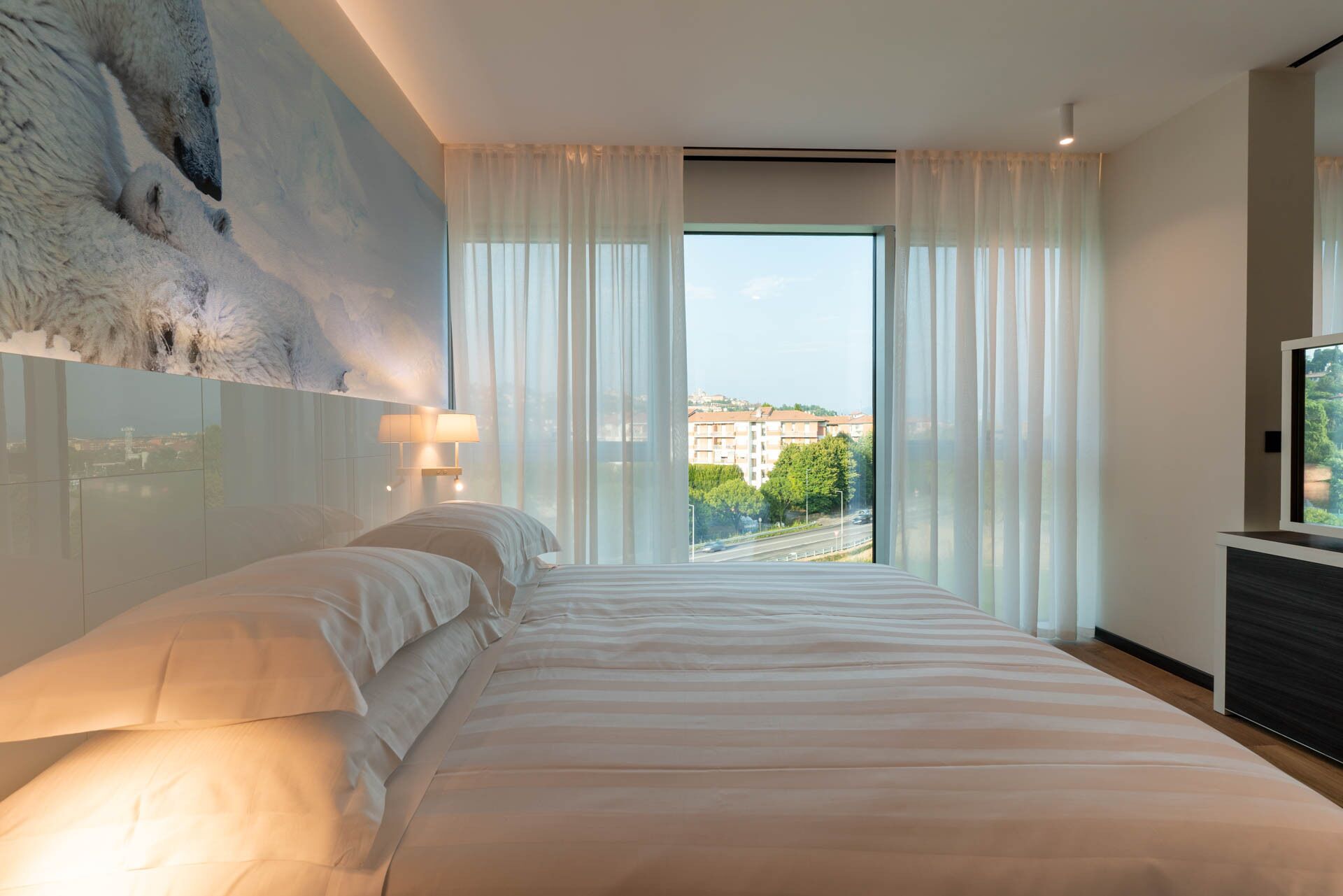 A modern hotel room with a large bed, a city view, and a mounted artwork on the wall.