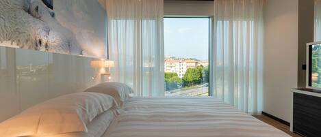 A modern hotel room with a large bed, a city view, and a mounted artwork on the wall.