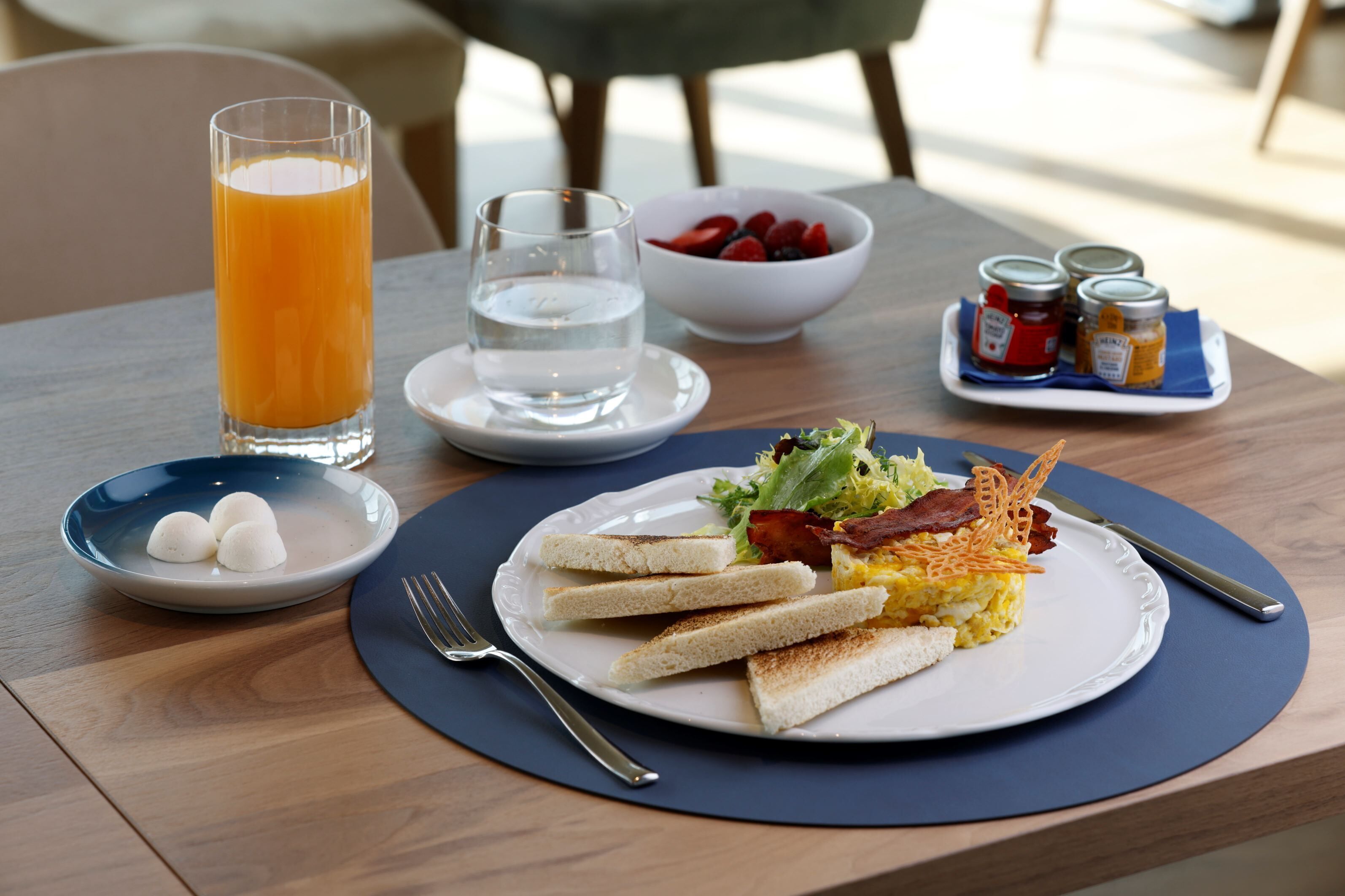 Un desayuno con tostadas, huevos revueltos, tocino y una ensalada en un plato, un vaso de jugo de naranja, un tazón de fresas y salsas en una bandeja.