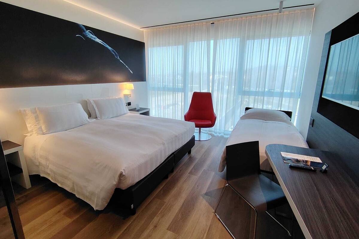 A hotel room with a large bed, a smaller bed, a desk with a laptop, a red chair, and a large window with sheer curtains.