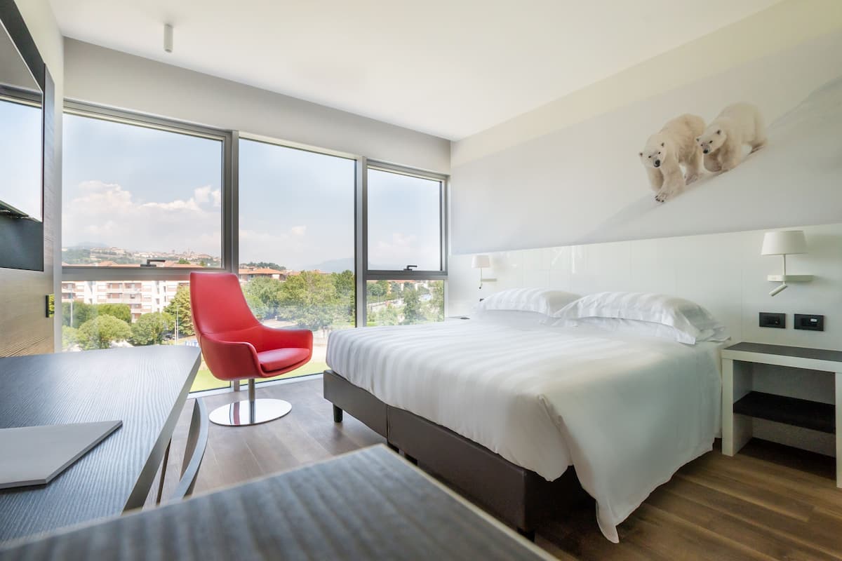 A modern hotel room with a large bed, a red chair, a desk, and a city view through large windows.