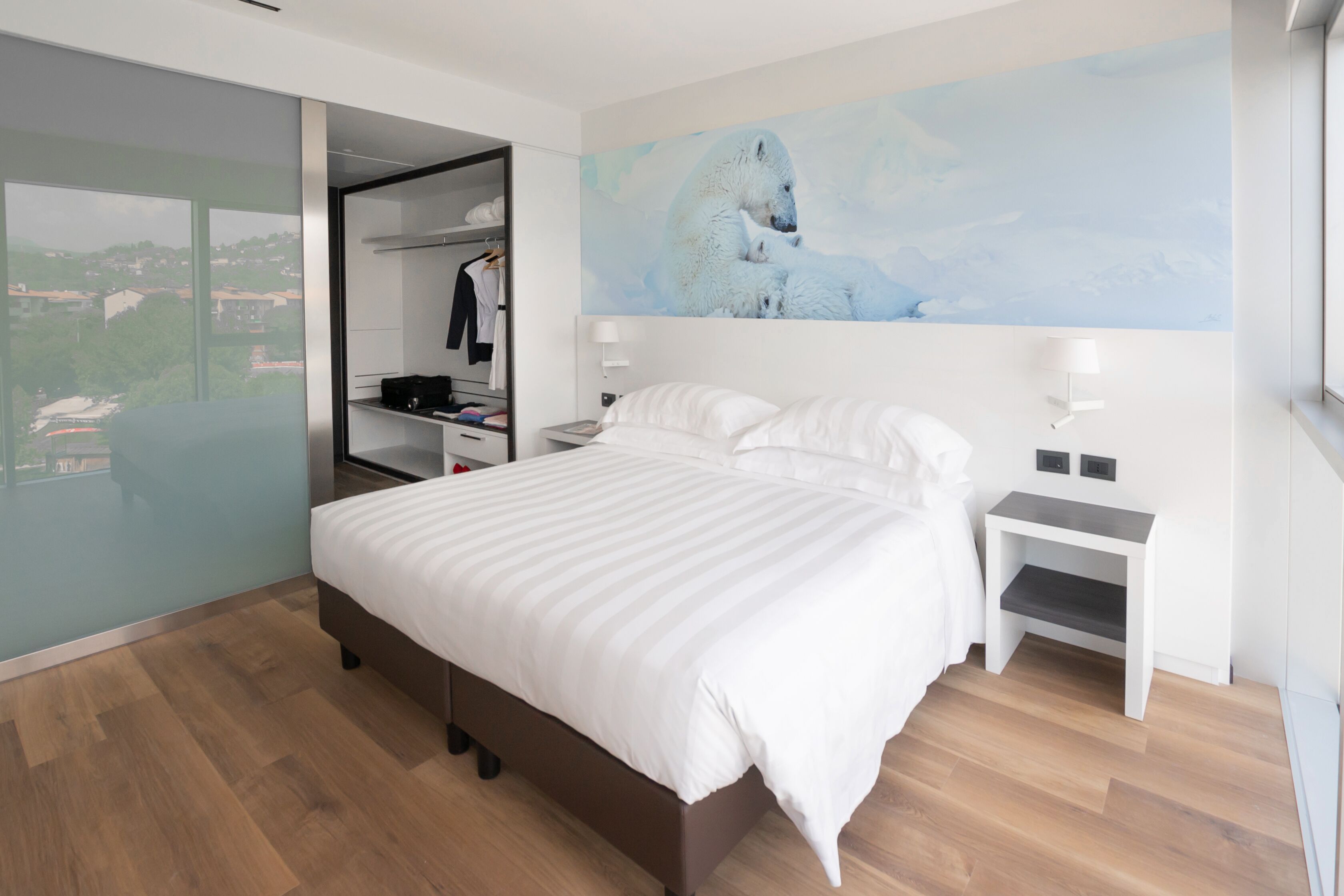 Una habitació d'hotel moderna amb un llit gran, un armari encastat, un quadre d'ossos polars i vista a un paisatge urbà.