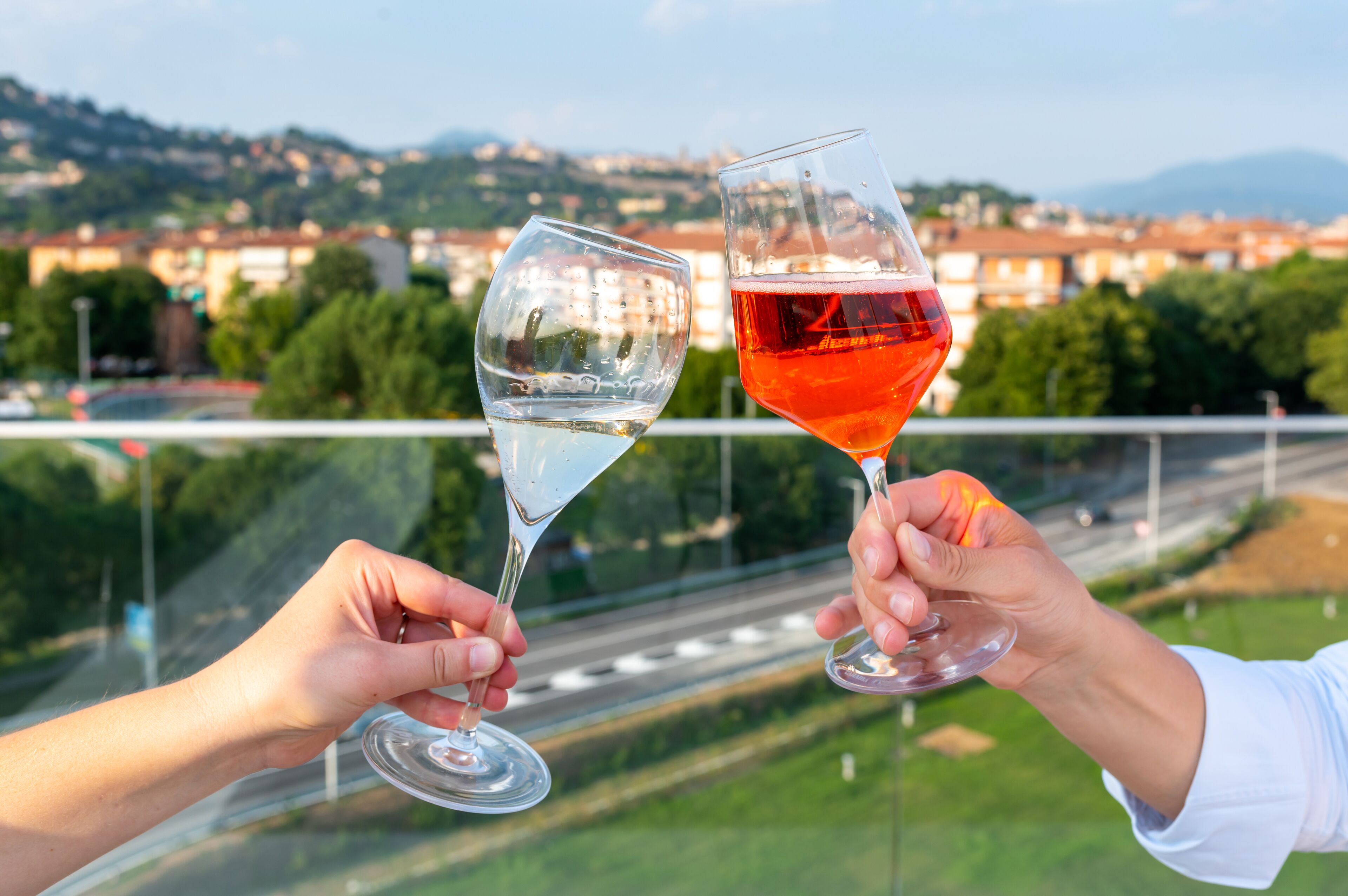 Two hands holding wine glasses with a scenic view of a city and mountains in the background.