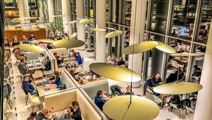 Uma sala de jantar moderna com várias mesas e cabines, luminárias pendentes grandes e vista da cidade através de amplas janelas.