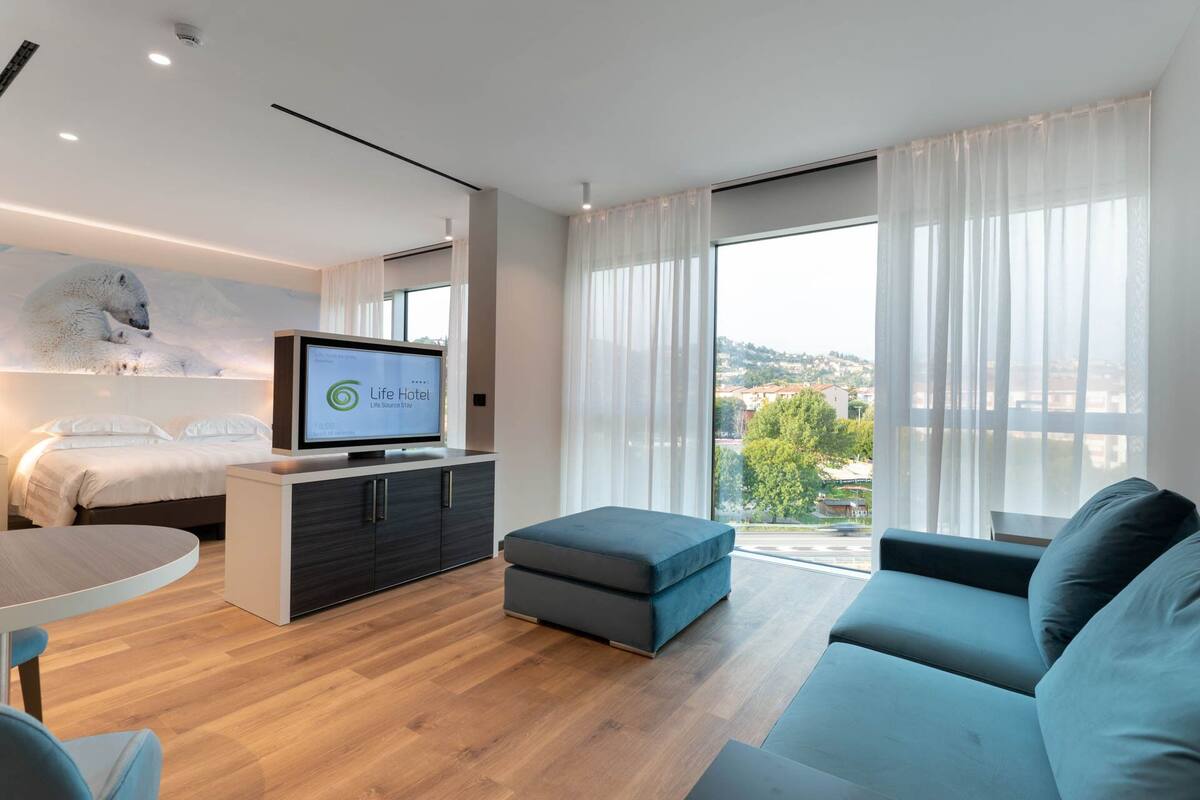 A modern hotel room with a bed, a TV, a sofa, and a balcony with a city view.