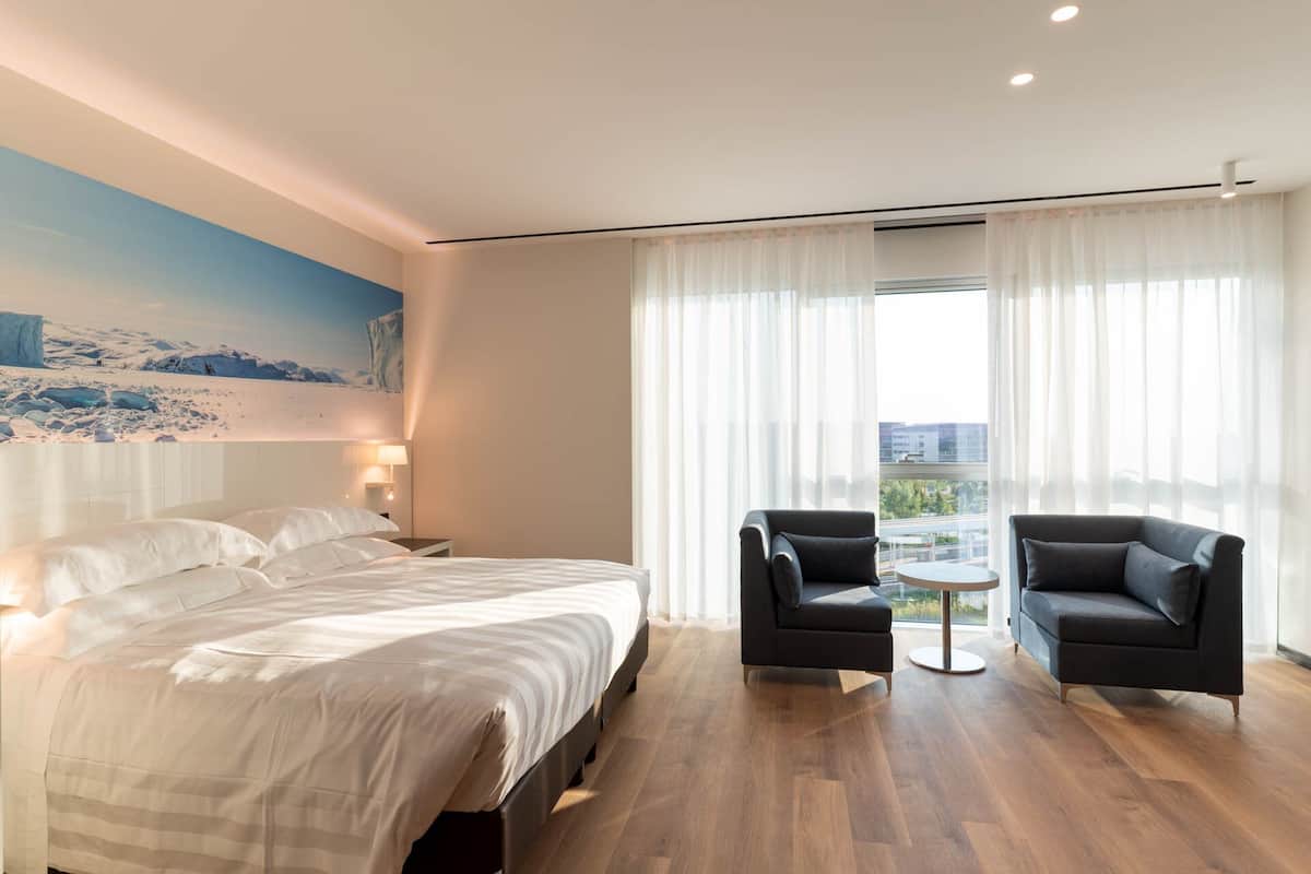 A hotel room with a large bed, two armchairs, a small table, and a city view through sheer curtains.
