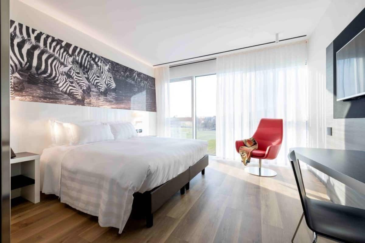 A modern hotel room with a large bed, a red chair, a flat-screen TV, and a zebra painting on the wall.