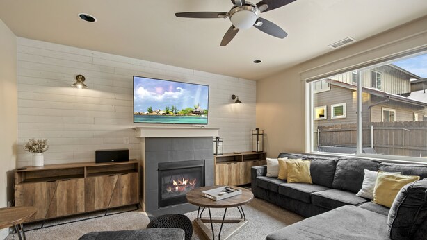 A modern living room with a fireplace, a flat-screen TV mounted above it, a ceiling fan, and a large window providing a view of the outside. - Pico in the Pines, pet friendly and cozy fireplace (Flagstaff)