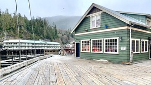 A wooden dock leading to a green building with a sloped roof, surrounded by a forested area and a misty mountain backdrop.