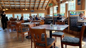 A spacious restaurant with wooden tables and chairs, large windows, and a high ceiling with exposed beams.