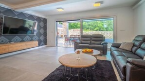 Ein modernes Wohnzimmer mit einem großen Fernseher, einem Sofa, einem Couchtisch mit Früchten und Blick ins Freie.