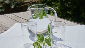 Un jarro transparente con hojas de menta sobre un mantel blanco, junto a dos vasos vacíos.