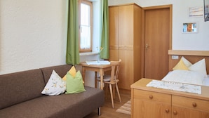 Una habitación acogedora con un armario de madera, un escritorio de madera con una silla, un sofá marrón con cojines estampados y una cama con edredón blanco.
