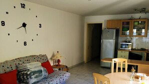 A living room with a sofa, a dining table with chairs, a kitchen with cabinets and appliances, and a wall clock. - Rental with swimming pool, close to beaches, at the foot of the maquis. (Biguglia)