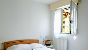 A bedroom with a wooden bed, a bedside table, a radiator, and a window with curtains. - Rental with swimming pool, close to beaches, at the foot of the maquis. (Biguglia)