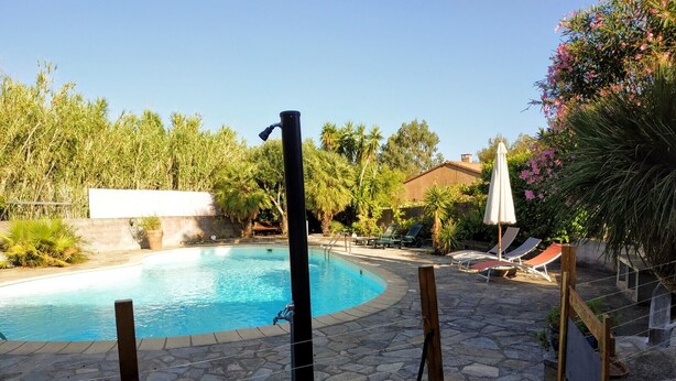 A swimming pool with a stone deck, surrounded by lounge chairs and umbrellas, with greenery and a building in the background. - Rental with swimming pool, close to beaches, at the foot of the maquis. (Biguglia)