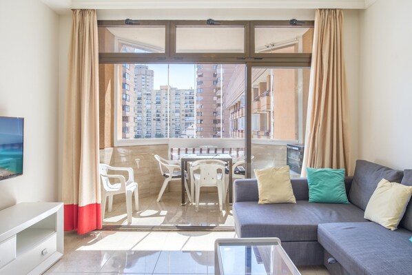Un balcón con vistas a edificios altos, un sofá gris con cojines, un mueble de televisión blanco y una mesa de vidrio.
