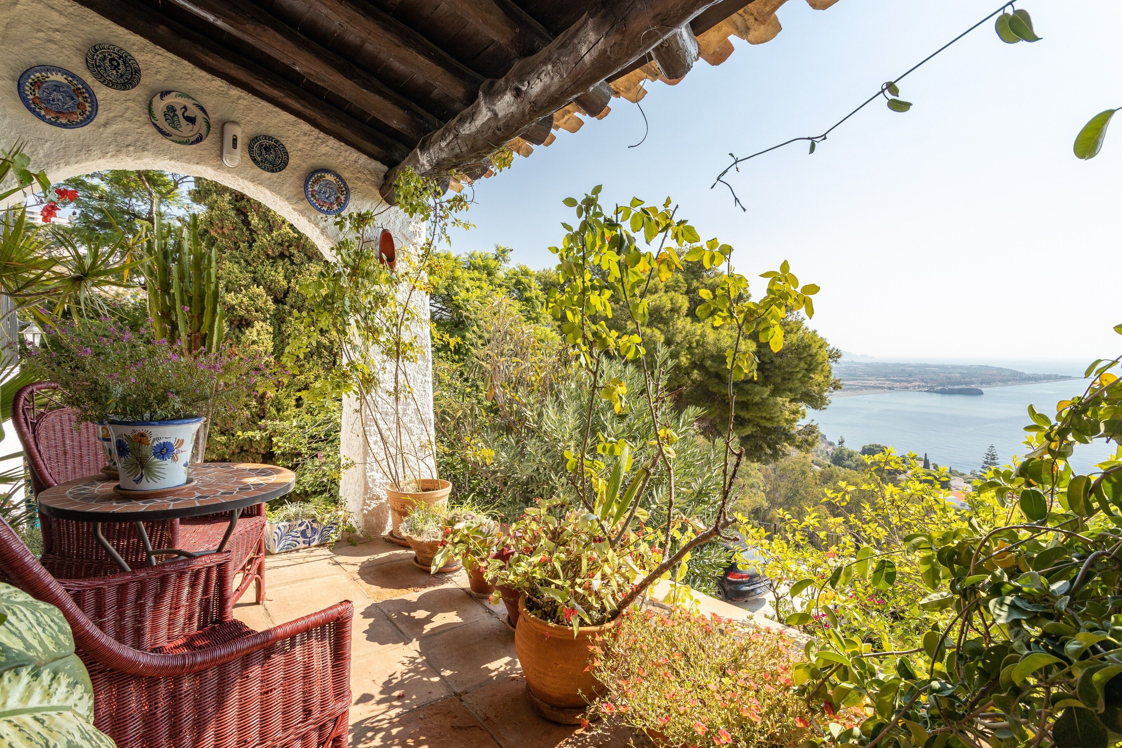 Eine Terrasse mit Korbmöbeln, Topfpflanzen und Blick auf das Meer.