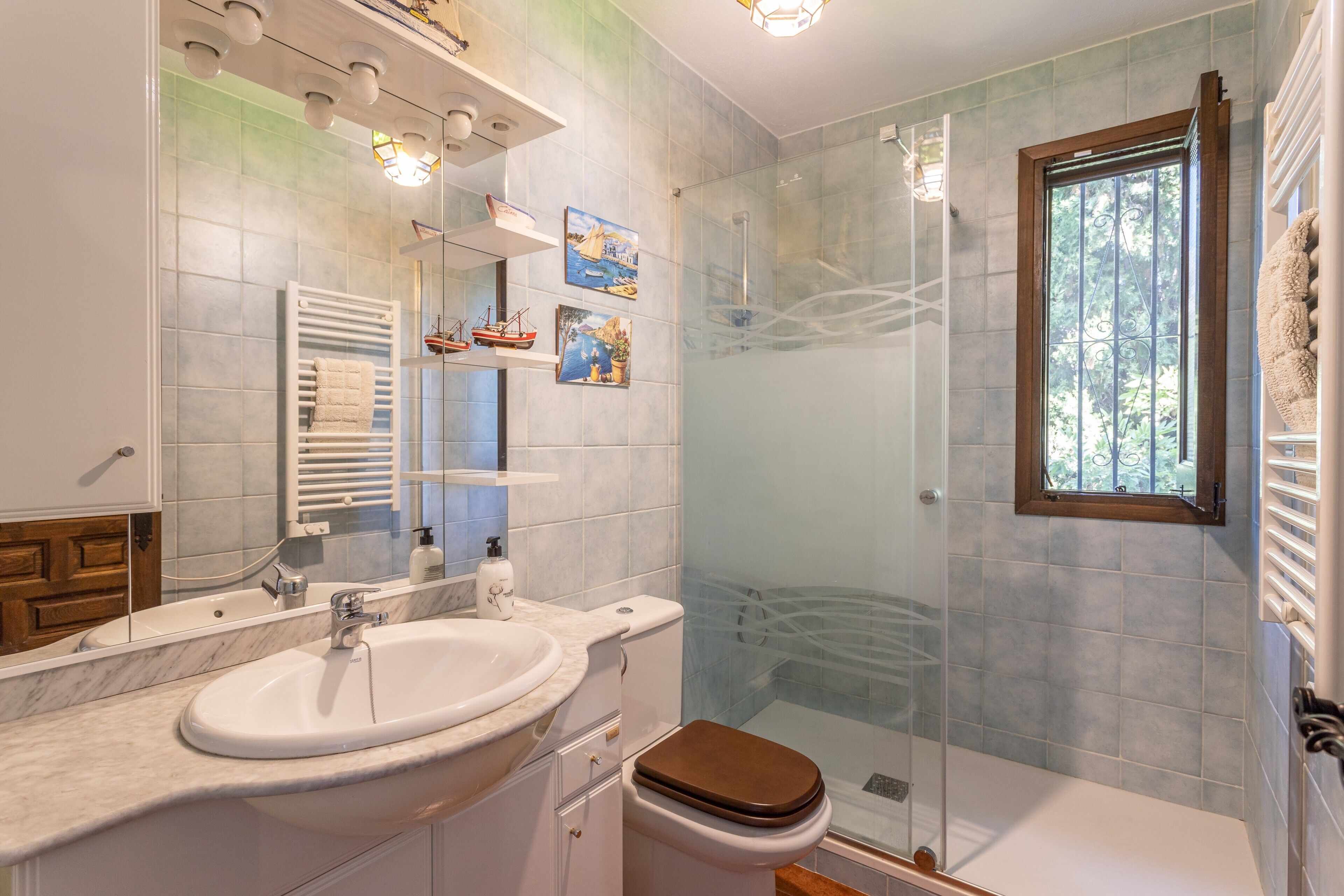 Un baño con cabina de ducha de vidrio, un lavabo blanco, inodoro y ventana con vistas a la vegetación.