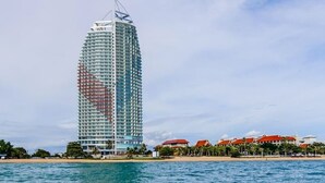 Un hotel alto y moderno con fachada curva, situado cerca de una playa con palmeras y un cielo azul claro.