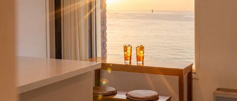 Une pièce avec vue sur la mer, une table avec deux verres remplis d’une boisson et une chaise avec des coussins.