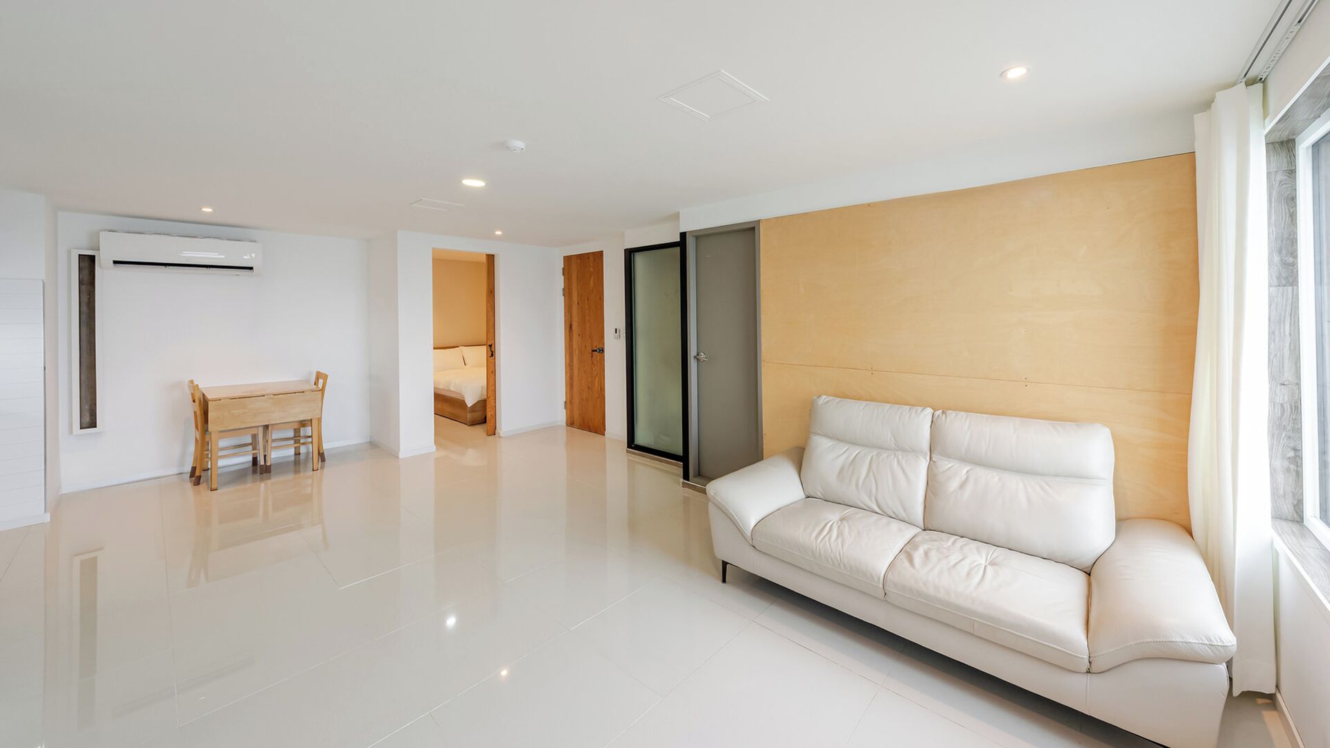 A modern living room with a white sofa, a wooden dining table, and a glass door leading to a bedroom.